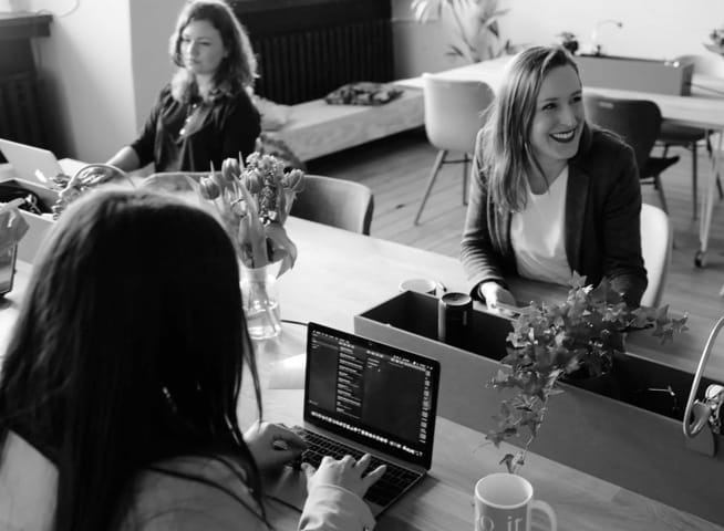 image of three women in a board meeting, sitted around a table, smiling, while pressing their laptops